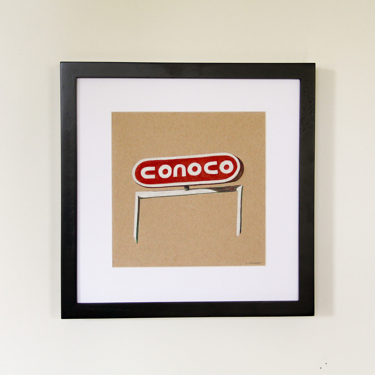 Framed artwork hung on white wall. Colored pencil drawing on tan toned paper of a red and white Conoco gas road sign with a 2 inch white matte around the art in a black wood frame. 